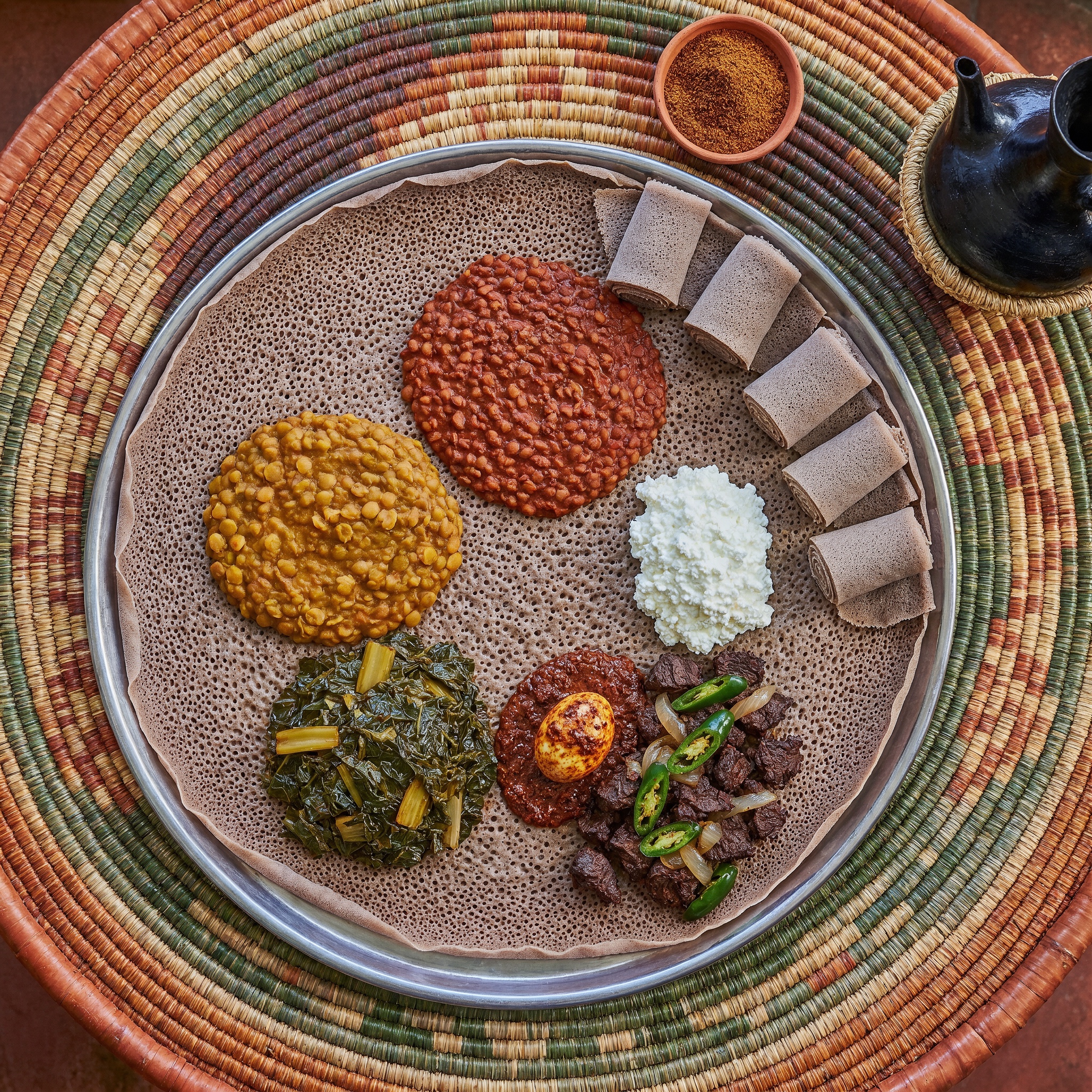 Overhead view of an Ethiopian injera platter topped with lentil, split-pea, collard, beef and egg dishes on a woven basket table.